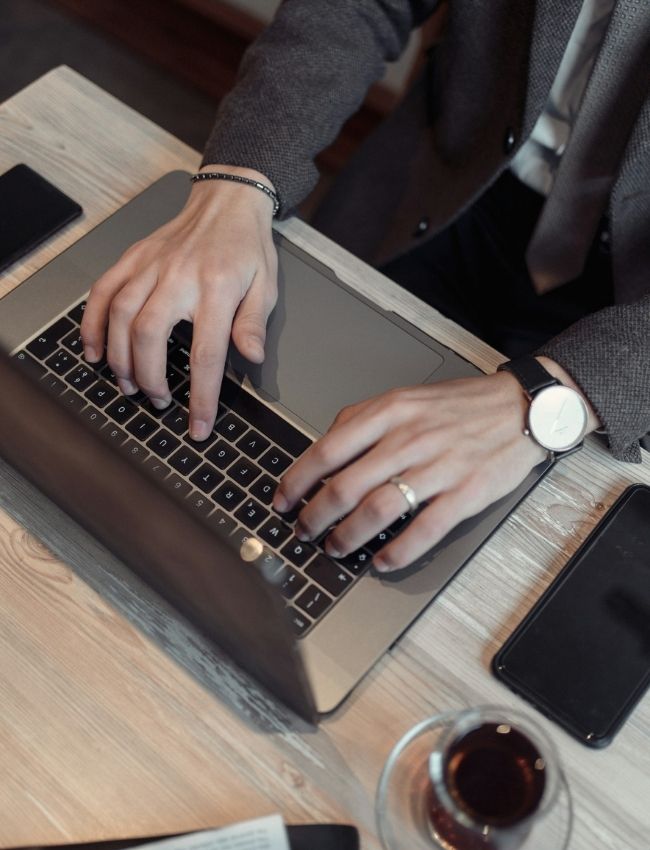 Person in Business-Kleidung tippt auf einem Laptop, während ein Smartphone und Kaffee daneben liegen